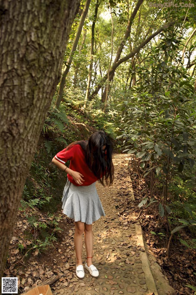A woman in a red shirt and gray skirt standing on a path in the woods.